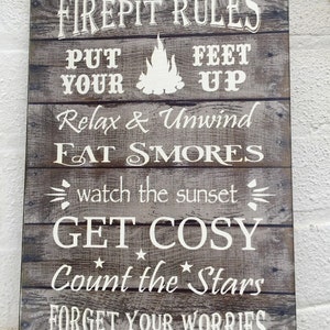 May include: A rustic wooden sign with white text that reads "Firepit Rules: Put Your Feet Up, Relax & Unwind, Eat Smores, Watch the Sunset, Get Cozy, Count the Stars, Forget Your Worries".
