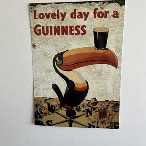 Puede incluir: Un letrero de estilo vintage con un tuc&aacute;n sosteniendo una pinta de Guinness en su pico. El texto "Lovely day for a Guinness" est&aacute; en letras rojas. El fondo muestra un paisaje urbano y una br&uacute;jula.