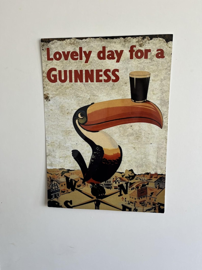 Puede incluir: Un letrero de estilo vintage con un tuc&aacute;n sosteniendo una pinta de Guinness en su pico. El texto "Lovely day for a Guinness" est&aacute; en letras rojas. El fondo muestra un paisaje urbano y una br&uacute;jula.