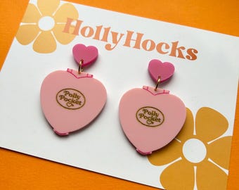 May include: A pair of pink heart-shaped earrings with a gold Polly Pocket logo. The earrings are on a white card with the text "Holly Hocks" in gold lettering.