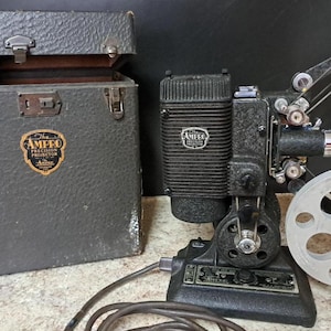 Könnte beinhalten: Ein antiker Ampro Präzisionsprojektor mit passendem Koffer. Der Projektor ist schwarz mit silbernen Akzenten und einer Filmrolle. Der Koffer ist dunkelgrau mit Metallverschlüssen und dem Ampro-Logo.