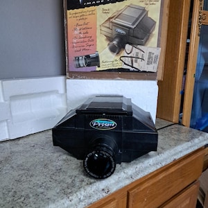 May include: A black Prism image projector with a large lens, sitting on a countertop. The box behind it reads "Super Prism Image Projector" and lists applications like fine art and illustrations.