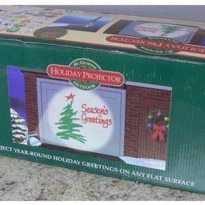 May include: A green cardboard box for a holiday projector. The box features a graphic of a projected Christmas scene with a tree and the words "Season's Greetings." The box also states "Project year-round holiday greetings on any flat surface."