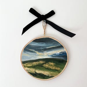May include: A round wooden ornament featuring a painted landscape scene of a green field with a blue sky and white clouds. The ornament is suspended from a black velvet ribbon with a crystal bead.