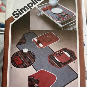 May include: A Simplicity 9794 sewing pattern envelope featuring illustrations of table linens including a long runner, rectangular placemats, a round placemat, and a solid red placemat, all displayed with plates, cutlery, and a teapot.