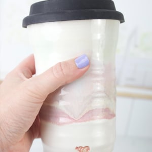 Könnte beinhalten: Ein Reisebecher aus Keramik mit weißer Glasur und weichen, horizontalen Farbverläufen in Rosa und Creme. Der Becher verfügt über einen schwarzen Silikondeckel und ein kleines, rotbraunes, herzförmiges Stempelmotiv am unteren Rand.