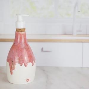 May include: A ceramic soap dispenser with a white pump and a cork stopper. The bottle has a white base, with a pink, dripped glaze. The dispenser is on a white countertop in a kitchen setting.