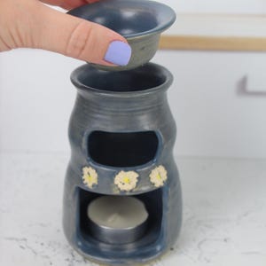 Puede incluir: Un quemador de aceite de cerámica azul con un cuenco superior extraíble y una cámara inferior que contiene una vela de té blanca. El exterior presenta tres pequeñas margaritas blancas pintadas a mano con centros amarillos.