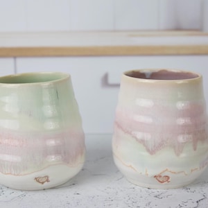 May include: Two ceramic tumblers with a rounded, handleless shape. The left tumbler features a pale green upper section transitioning into a soft pink and white base. The right tumbler has a pale pink upper section fading into white.