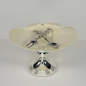 Könnte beinhalten: Eine dekorative Servierschale mit einer großen, cremefarbenen Muschelschale. Die Schale wird von einem silberfarbenen Sockel getragen. Zwei silberfarbene Dekorationselemente sind an der Schale befestigt.