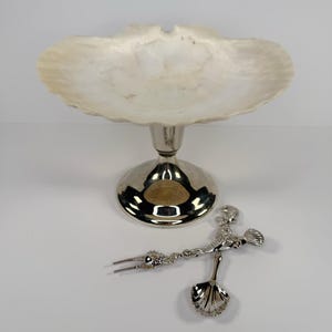 May include: A decorative serving dish with a white, shell-shaped bowl and a silver-coloured pedestal base. A silver-coloured serving fork and spoon set with seashell and floral details rests nearby. The items are on a white surface.