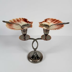 May include: A decorative, two-part serving dish with a silver-toned metal base. Each side features a shell-shaped dish in cream and reddish-brown hues, with a small spoon resting inside. The base has an ornate design.