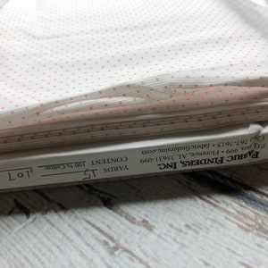 May include: A roll of white fabric with a repeating pattern of small pink polka dots. The fabric is labeled as 100% cotton and contains 15 yards. The label also includes the manufacturer's information.