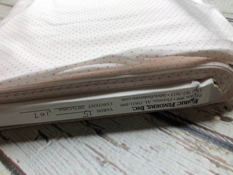 May include: A roll of white fabric with a repeating pattern of small pink polka dots. The fabric is labeled as 100% cotton and contains 15 yards. The label also includes the manufacturer's information.