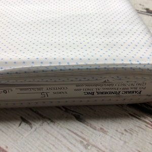 May include: A roll of white cotton fabric featuring a pattern of small, evenly spaced light blue dots. The fabric is labelled with the manufacturer's details, including the content, which is 100% cotton, and the length, which is 13.7 metres.