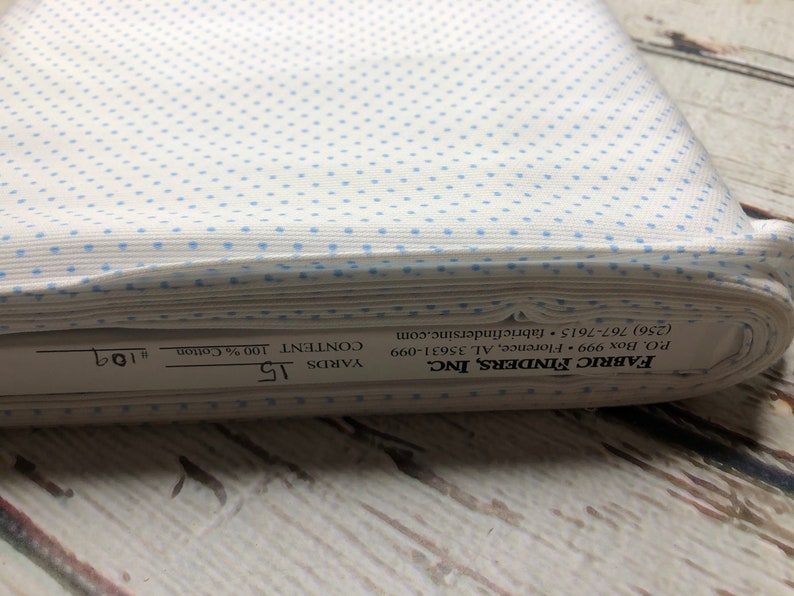 May include: A roll of white cotton fabric featuring a pattern of small, evenly spaced light blue dots. The fabric is labelled with the manufacturer's details, including the content, which is 100% cotton, and the length, which is 13.7 metres.