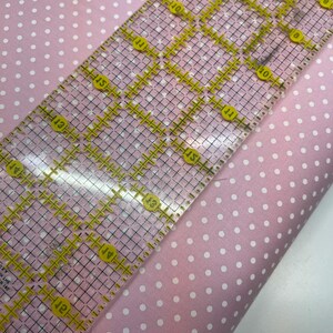 May include: A light pink fabric featuring a repeating pattern of small white polka dots. A clear acrylic quilting ruler with yellow grid markings and numbers is placed diagonally across the fabric, showing a width of approximately 6 inches.