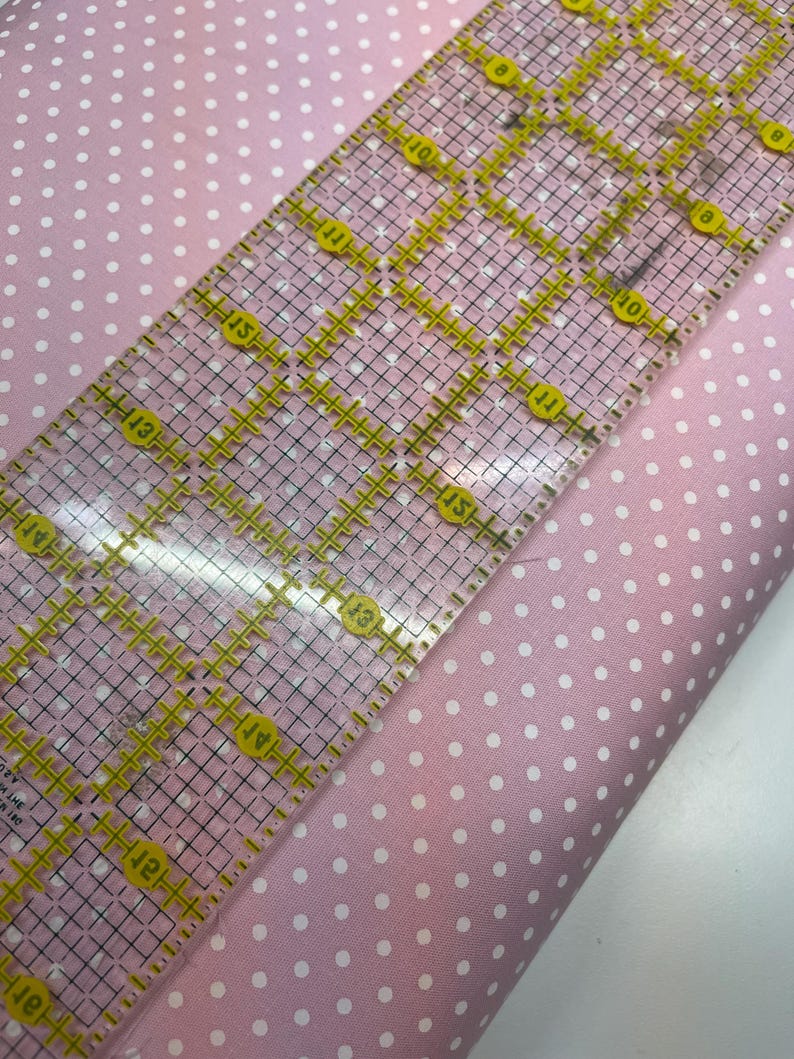 May include: A light pink fabric featuring a repeating pattern of small white polka dots. A clear acrylic quilting ruler with yellow grid markings and numbers is placed diagonally across the fabric, showing a width of approximately 6 inches.