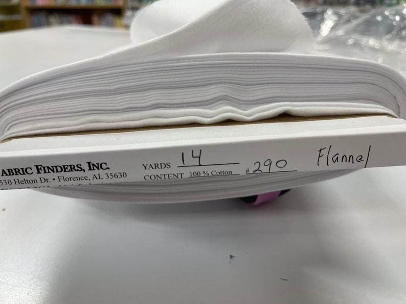 Op de afbeelding: Witte flanellen stof gevouwen in een stapel. Het label vermeldt "Fabric Finders, Inc." "530 Helton Dr. Florence, AL 35630" "Yards 14" "Content 100% Cotton" "#290 Flannel"