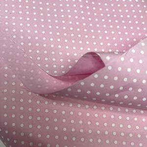 May include: A light pink fabric featuring a repeating pattern of small, evenly spaced white polka dots. The material is draped to show its texture and soft folds, highlighting the consistent contrast between the pink background and white dots.