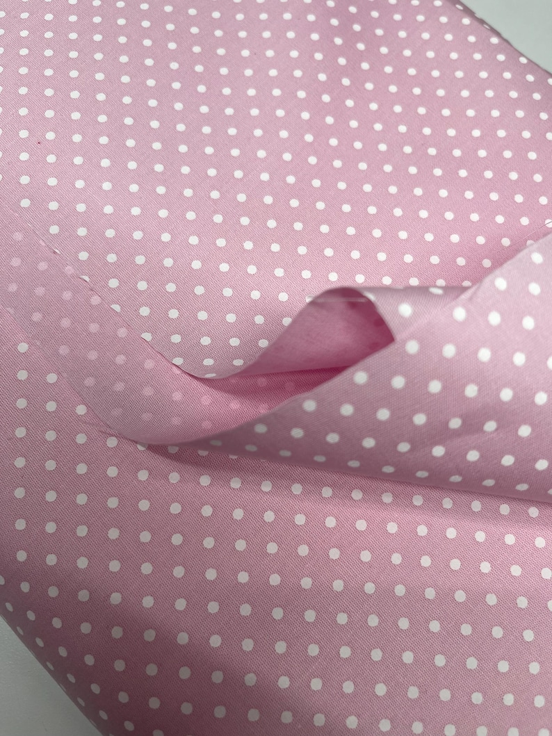May include: A light pink fabric featuring a repeating pattern of small, evenly spaced white polka dots. The material is draped to show its texture and soft folds, highlighting the consistent contrast between the pink background and white dots.