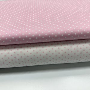 May include: Two folded layers of fabric featuring a classic polka dot pattern. The top layer is light pink with small white dots, while the bottom layer is white with small light pink dots. The fabric is smooth and neatly stacked in horizontal folds.