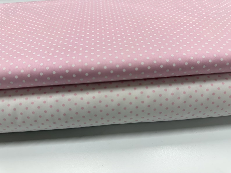 May include: Two folded layers of fabric featuring a classic polka dot pattern. The top layer is light pink with small white dots, while the bottom layer is white with small light pink dots. The fabric is smooth and neatly stacked in horizontal folds.