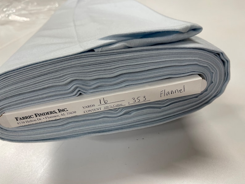 May include: A roll of light blue flannel fabric with a white label that reads "FABRIC FINDERS, INC. 4530 Helton Dr. Florence, AL 35630 YARDS 16 CONTENT 100% Cotton 353 Flannel"