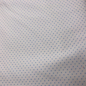 May include: White fabric with a repeating pattern of small, light blue polka dots. The fabric has a textured appearance and is folded, showing the pattern across the surface. The background is a light-colored wooden surface.