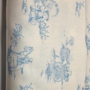May include: Off-white fabric with a repeating blue toile pattern featuring teddy bears, trees, and carriages. The design is reminiscent of classic nursery decor, with a vintage aesthetic. The fabric appears to be cotton or linen.
