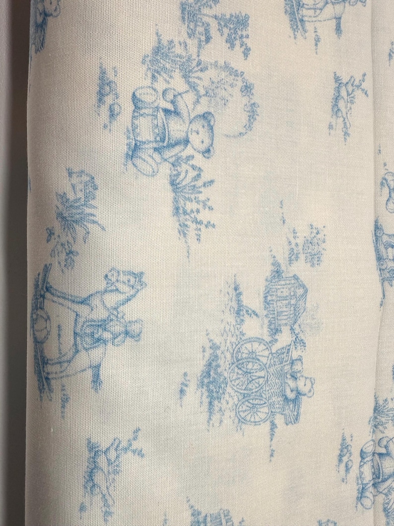 May include: Off-white fabric with a repeating blue toile pattern featuring teddy bears, trees, and carriages. The design is reminiscent of classic nursery decor, with a vintage aesthetic. The fabric appears to be cotton or linen.