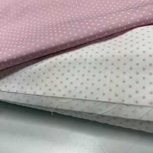 May include: Two folded pieces of fabric featuring a polka dot pattern. The top layer is light pink with small white dots, while the bottom layer is white with small light pink dots. The fabric edges show visible loose threads and a woven texture.