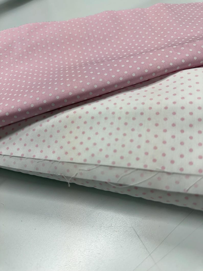May include: Two folded pieces of fabric featuring a polka dot pattern. The top layer is light pink with small white dots, while the bottom layer is white with small light pink dots. The fabric edges show visible loose threads and a woven texture.