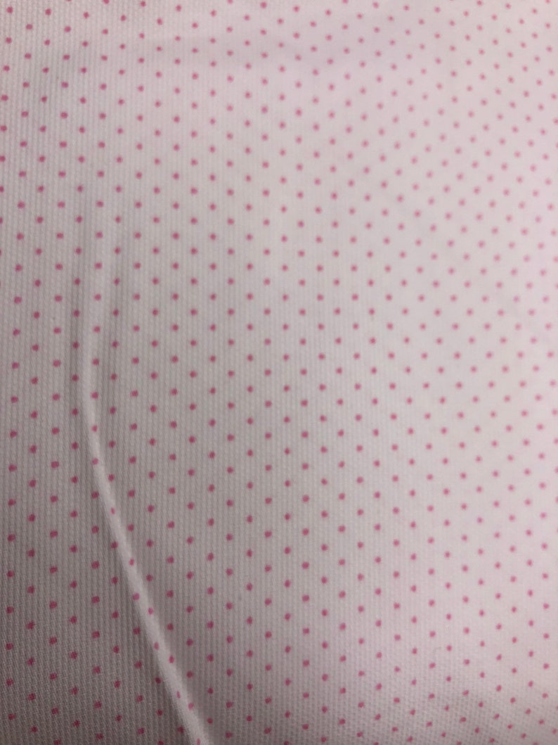 May include: Close-up of a white fabric with a repeating pattern of small, evenly spaced pink polka dots. The fabric has a subtle texture, and the dots are consistent in size and color, creating a delicate and classic design.