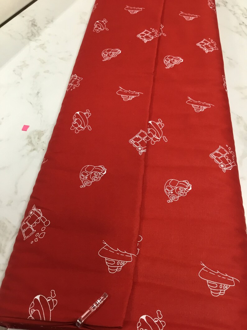 May include: Red fabric with a white repeating pattern of small cars. The fabric is folded in half.
