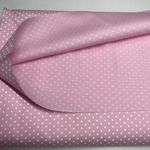 May include: A folded piece of light pink fabric featuring a repeating pattern of small white polka dots. The material is layered, showing the texture of the textile with a soft, matte finish and a consistent, evenly spaced dot design throughout.
