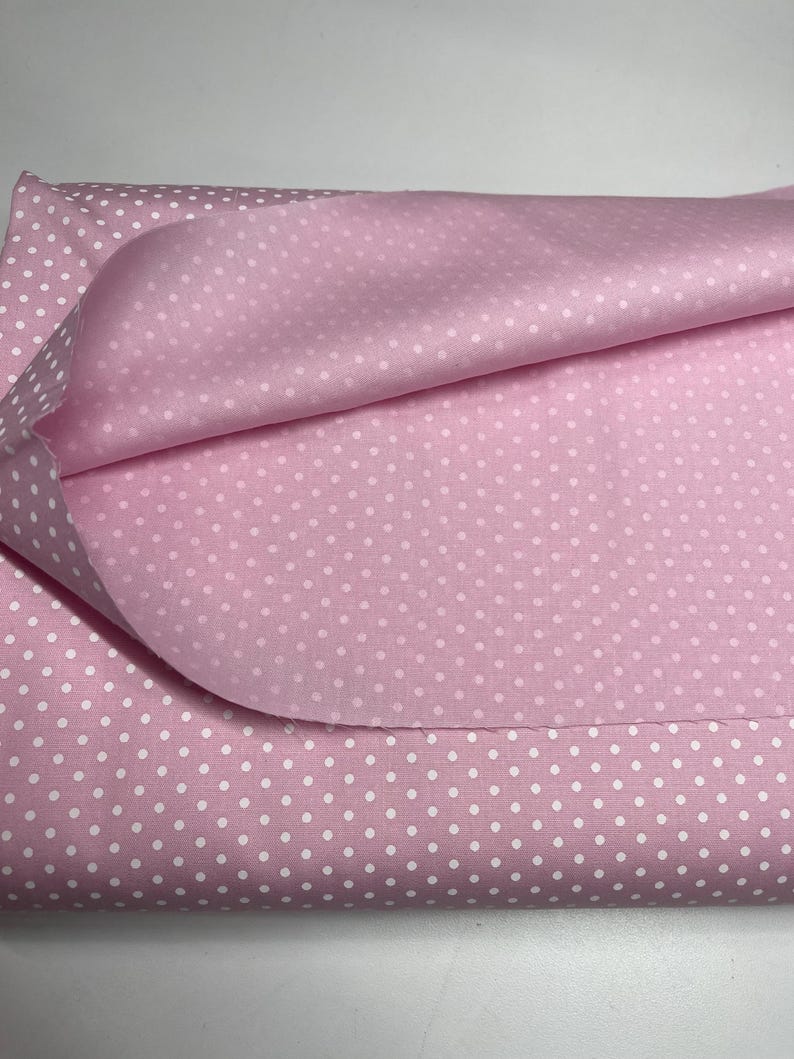 May include: A folded piece of light pink fabric featuring a repeating pattern of small white polka dots. The material is layered, showing the texture of the textile with a soft, matte finish and a consistent, evenly spaced dot design throughout.
