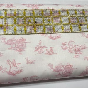 May include: White fabric with a pink toile pattern featuring teddy bears, carriages, and rocking horses. A clear ruler with yellow markings is placed on top of the fabric. The fabric is likely used for sewing or crafting projects.