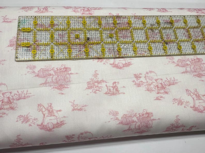 May include: White fabric with a pink toile pattern featuring teddy bears, carriages, and rocking horses. A clear ruler with yellow markings is placed on top of the fabric. The fabric is likely used for sewing or crafting projects.
