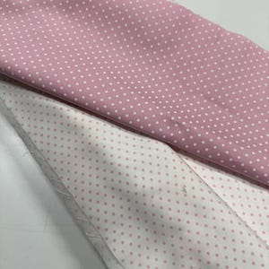May include: A folded length of fabric featuring two distinct patterns of white polka dots on a pink background and pink polka dots on a white background. The textile is layered, showing the contrast between the two colorways of the dotted print.