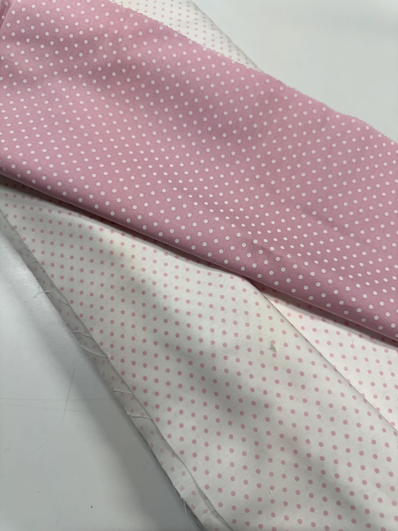 May include: A folded length of fabric featuring two distinct patterns of white polka dots on a pink background and pink polka dots on a white background. The textile is layered, showing the contrast between the two colorways of the dotted print.