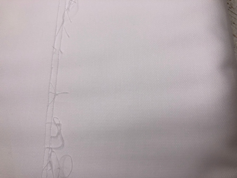 May include: Close-up of white fabric with a visible seam running vertically down the center. The fabric has a textured appearance, and loose threads are present along the seam's edge. The image highlights the texture and construction of the material.