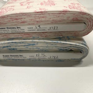 May include: Two rolls of fabric, one with a red and white toile pattern and the other with a blue and white toile pattern. The fabric is 100% cotton. The rolls have labels with the company name and address.
