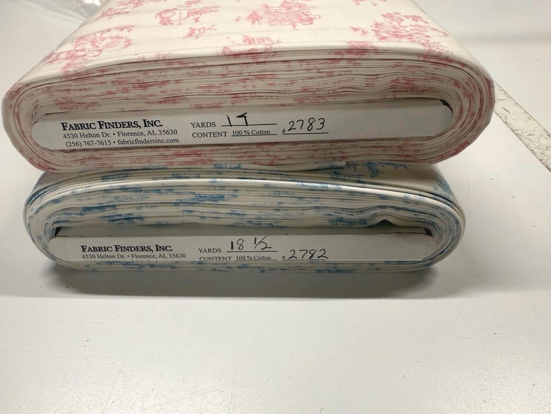 May include: Two rolls of fabric, one with a red and white toile pattern and the other with a blue and white toile pattern. The fabric is 100% cotton. The rolls have labels with the company name and address.