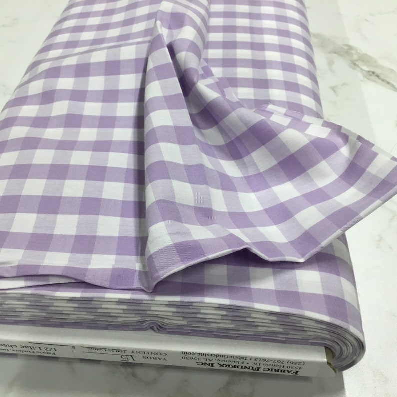 May include: A roll of white and lilac gingham fabric. The fabric is folded and the label is visible. The label reads "Fabric Finders, Inc. 4530 Helion Dr. Florence, AL 35630 (256) 767-7615 fabricfindersinc.com VARDS 15 CONTENTS 100% cotton 1/2 Lilac check"