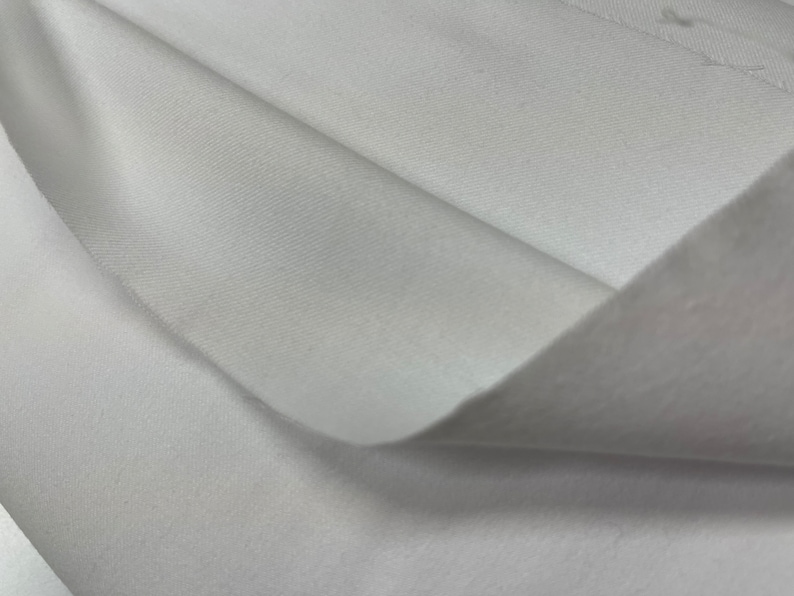 Op de afbeelding: Witte stof met een gladde, zijdeachtige textuur. De stof is doormidden gevouwen, waardoor de binnen- en buitenkant van het materiaal zichtbaar is.