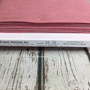 May include: A stack of red and white gingham fabric with a label that reads "Fabric Finders, Inc. Berry Micro Check 100% Cotton 18 5/8 yards".