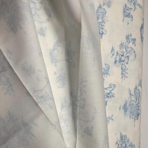 May include: White fabric with a blue toile pattern. The fabric is draped, showing the texture and design. The pattern features detailed illustrations of buildings, trees, and figures in a classic, elegant style.