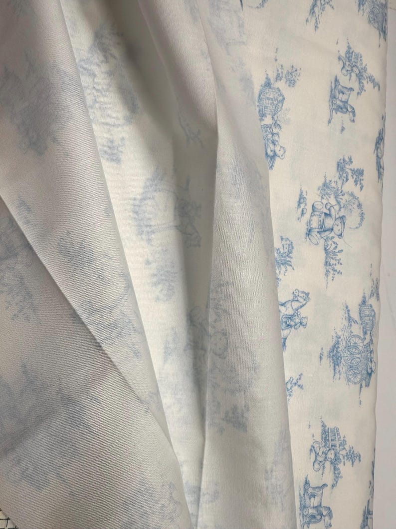 May include: White fabric with a blue toile pattern. The fabric is draped, showing the texture and design. The pattern features detailed illustrations of buildings, trees, and figures in a classic, elegant style.