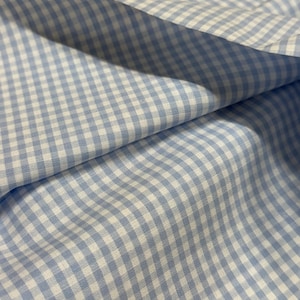 May include: Close-up of a light blue and white gingham fabric. The fabric is folded, showing the texture and pattern of the woven material. The pattern is a classic checkered design, with small squares of light blue and white.